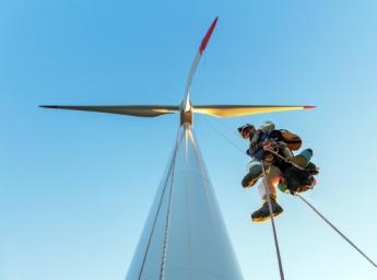 Low angle view of Rope Access Technician, industrial climber hanging on rope on wind turbine illuminated by sunset and looking into camera with shoe covers.